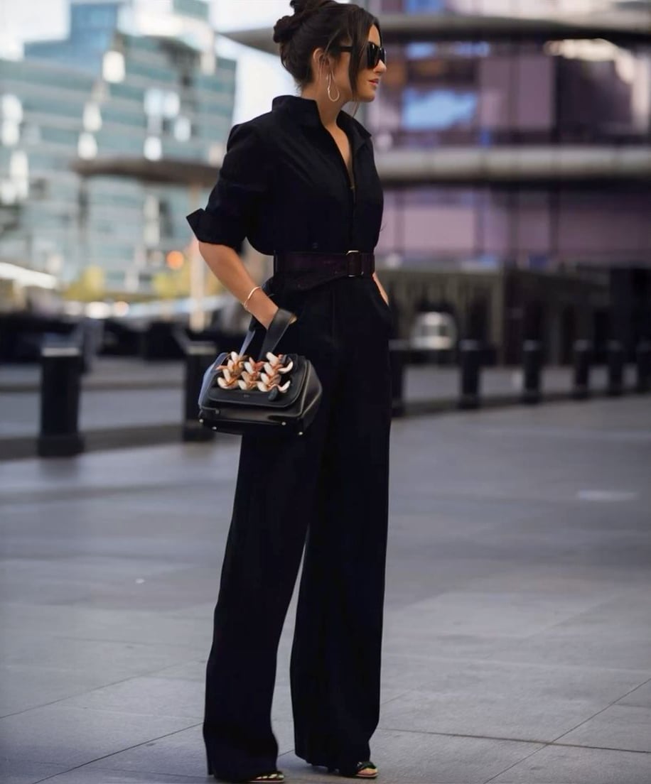 A woman wearing a stylish black jumpsuit, black gloves, and sunglasses stands outdoors in an urban setting, holding a black handbag with a chain detail. Modern buildings are blurred in the background.