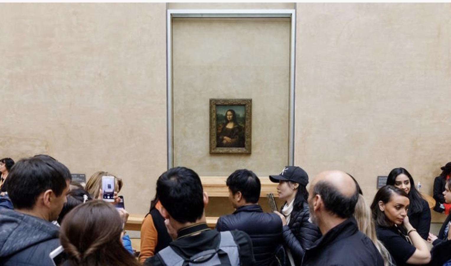 A crowd of people stands in front of the Mona Lisa painting, which is displayed on a beige wall in a museum. Some visitors are taking photos while others look at the artwork.