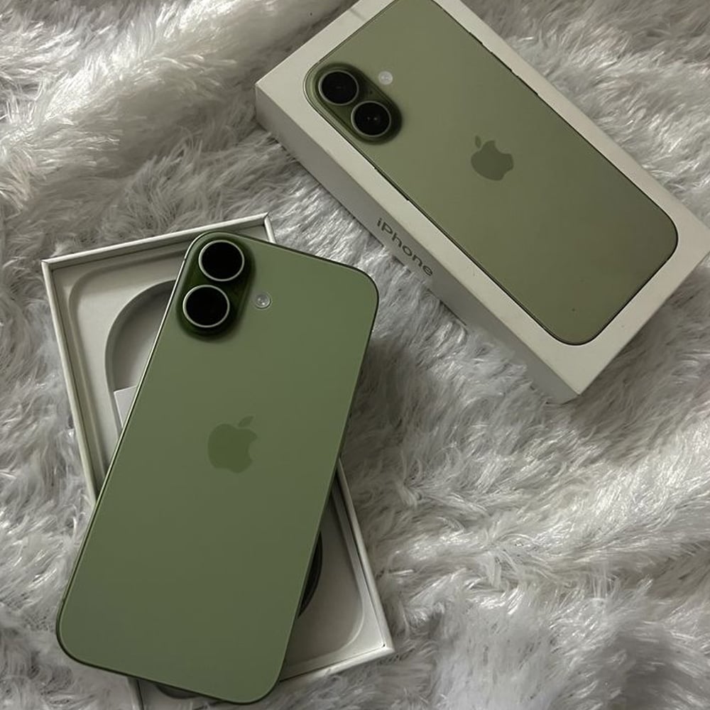 Two green iPhones and their white boxes are placed on a fluffy, light grey surface. One phone is inside an open box, and the other rests on top of a closed box. Both phones have dual rear cameras.