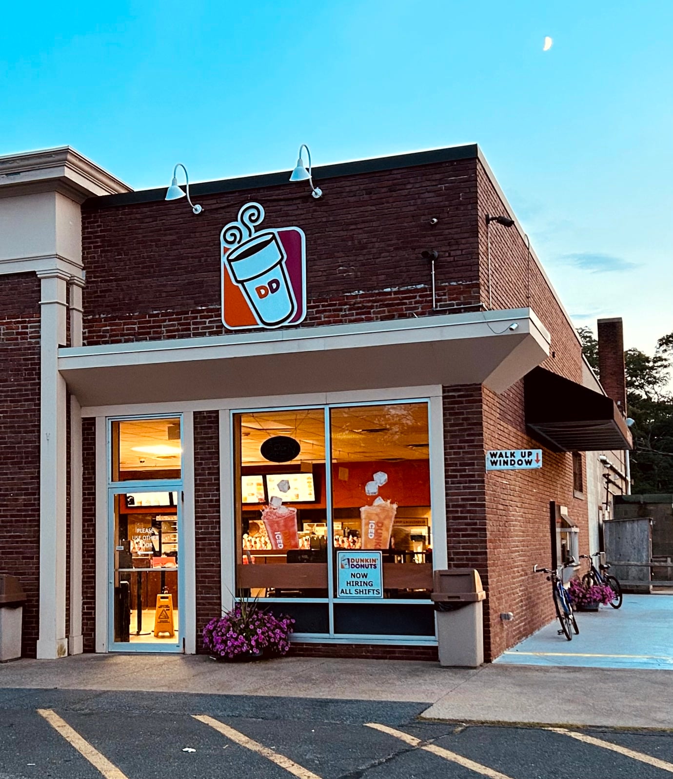A Dunkin’ Donuts store with large front windows, a brick exterior, and a walk-up window sign. The inside lights are on, and there are pink flowers and a bicycle outside. The DD coffee cup sign is above the entrance.
