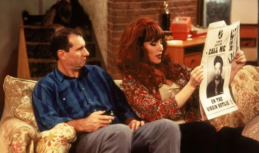 A man and a woman sit on a patterned couch; the woman, with red hair, is reading a newspaper with a large advertisement while the man looks at her. The scene appears to be from a sitcom set in a living room.