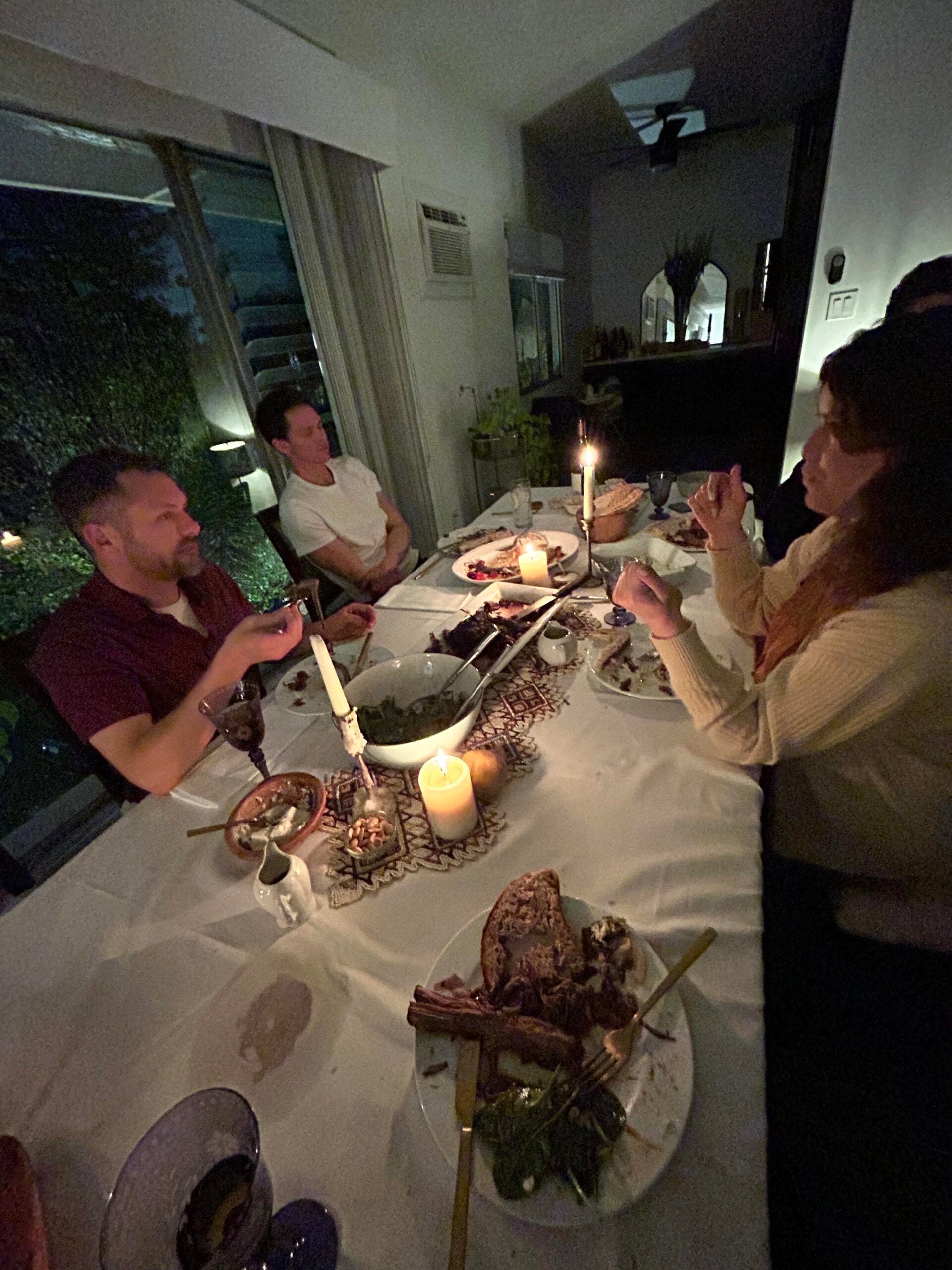 Four people sit around a candlelit dinner table at night, sharing food and conversation. Plates of food, utensils, and lit candles are spread across the white tablecloth. The setting is cozy and intimate.