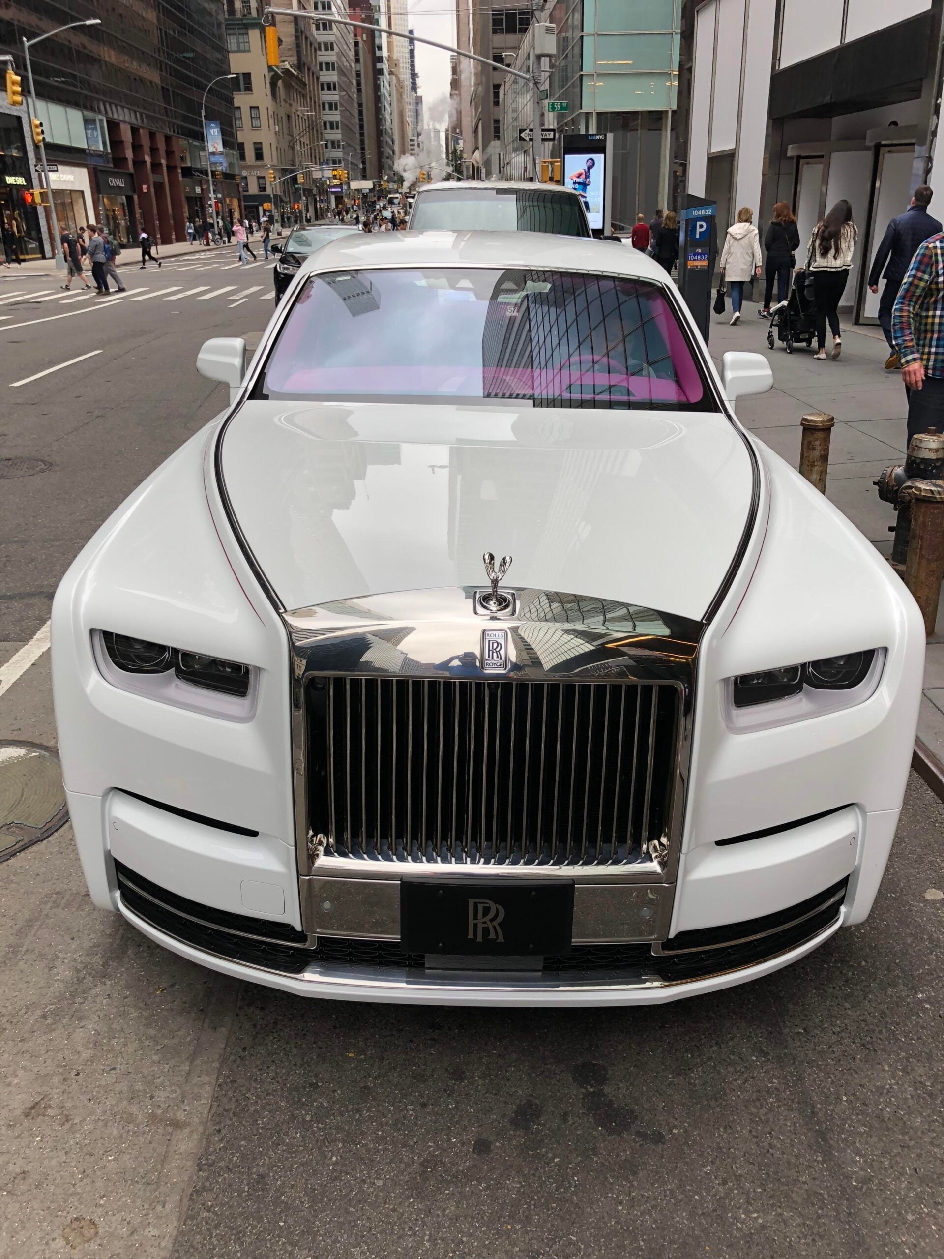 A white Rolls-Royce with a shiny chrome grille and hood ornament is parked on a city street, surrounded by tall buildings and people walking on the sidewalk.