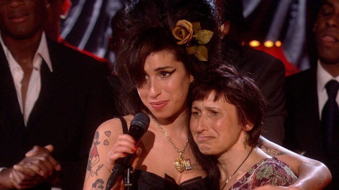 A woman with dark hair and tattoos, wearing a gold rose hair accessory, hugs another woman with short hair on stage. Both appear emotional, with people standing in the background.