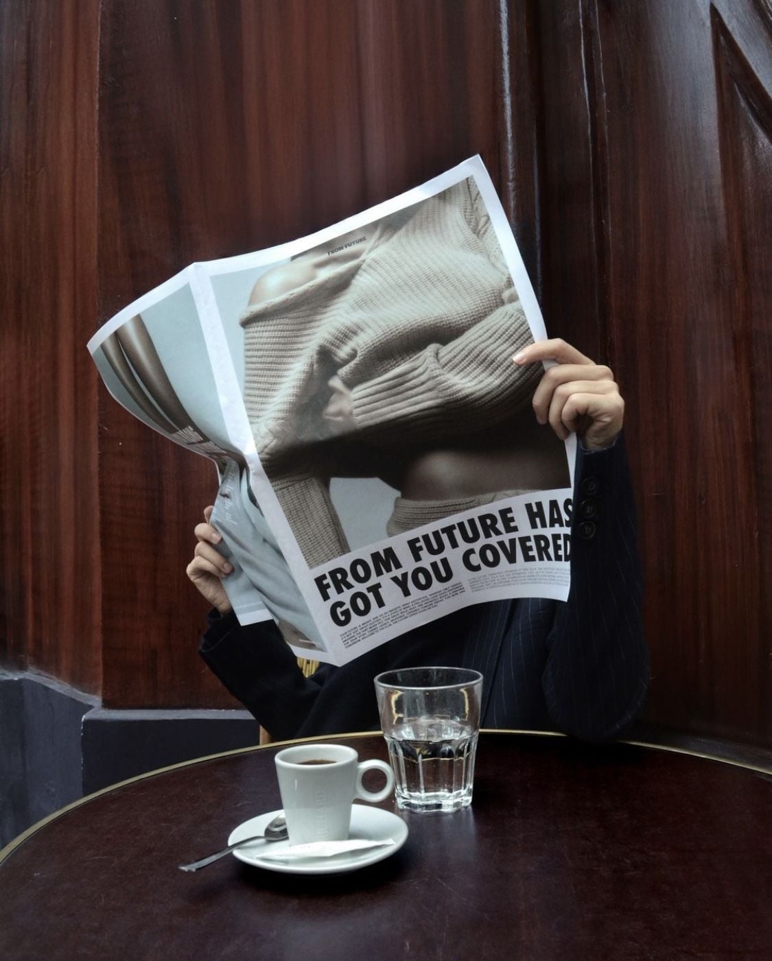 A person sits at a round table with a cup of espresso and a glass of water, holding up a large newspaper that covers their face. The headline reads “FROM FUTURE HAS GOT YOU COVERED.”