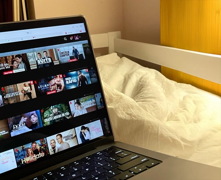 A laptop displaying a streaming service sits on a bed with white bedding in a cozy, softly lit bedroom. The screen shows various TV shows and movies to choose from.