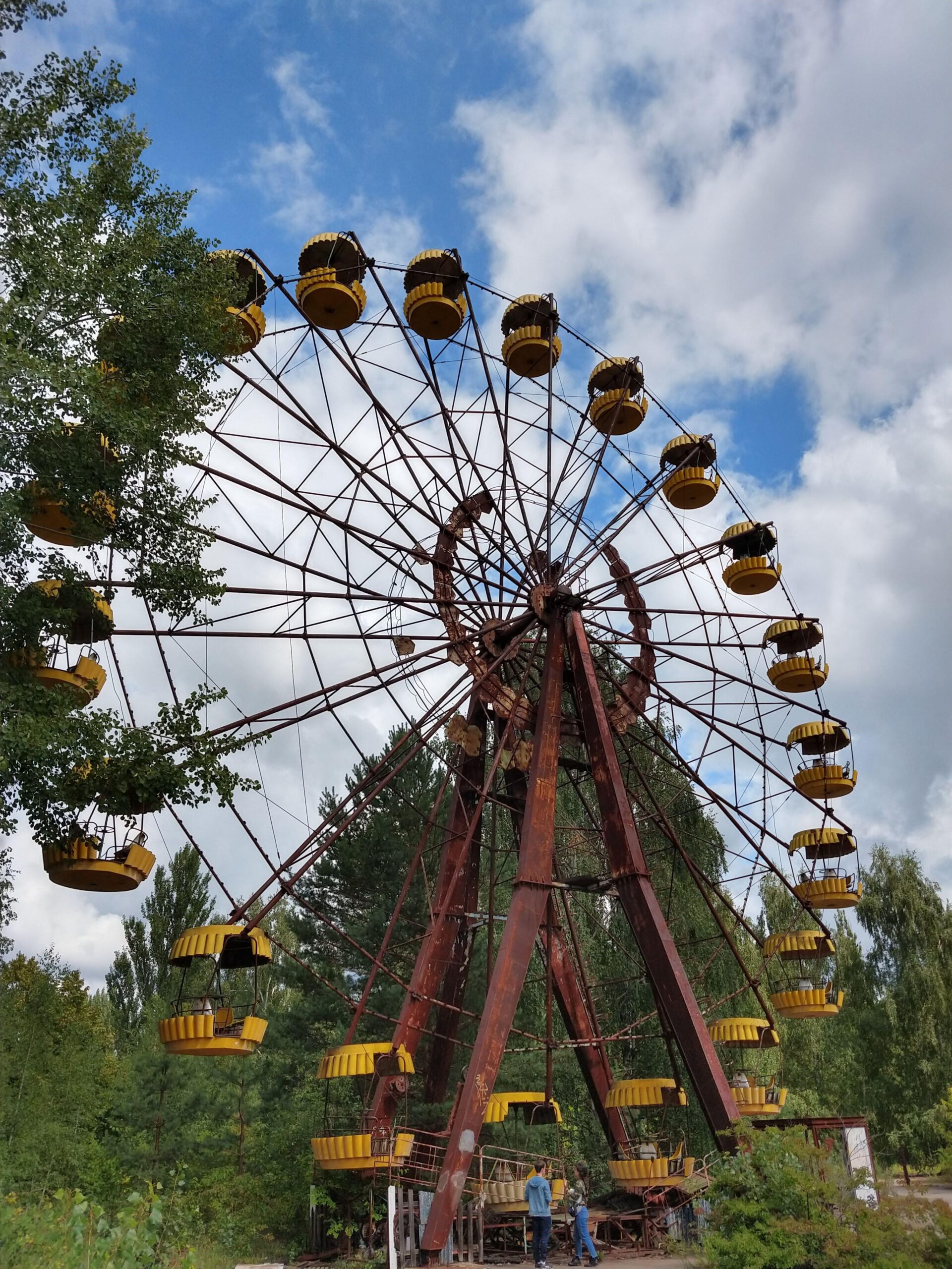 A large, rusted Ferris wheel with yellow passenger cars stands abandoned among overgrown greenery under a partly cloudy sky. Two people are seen near its base.