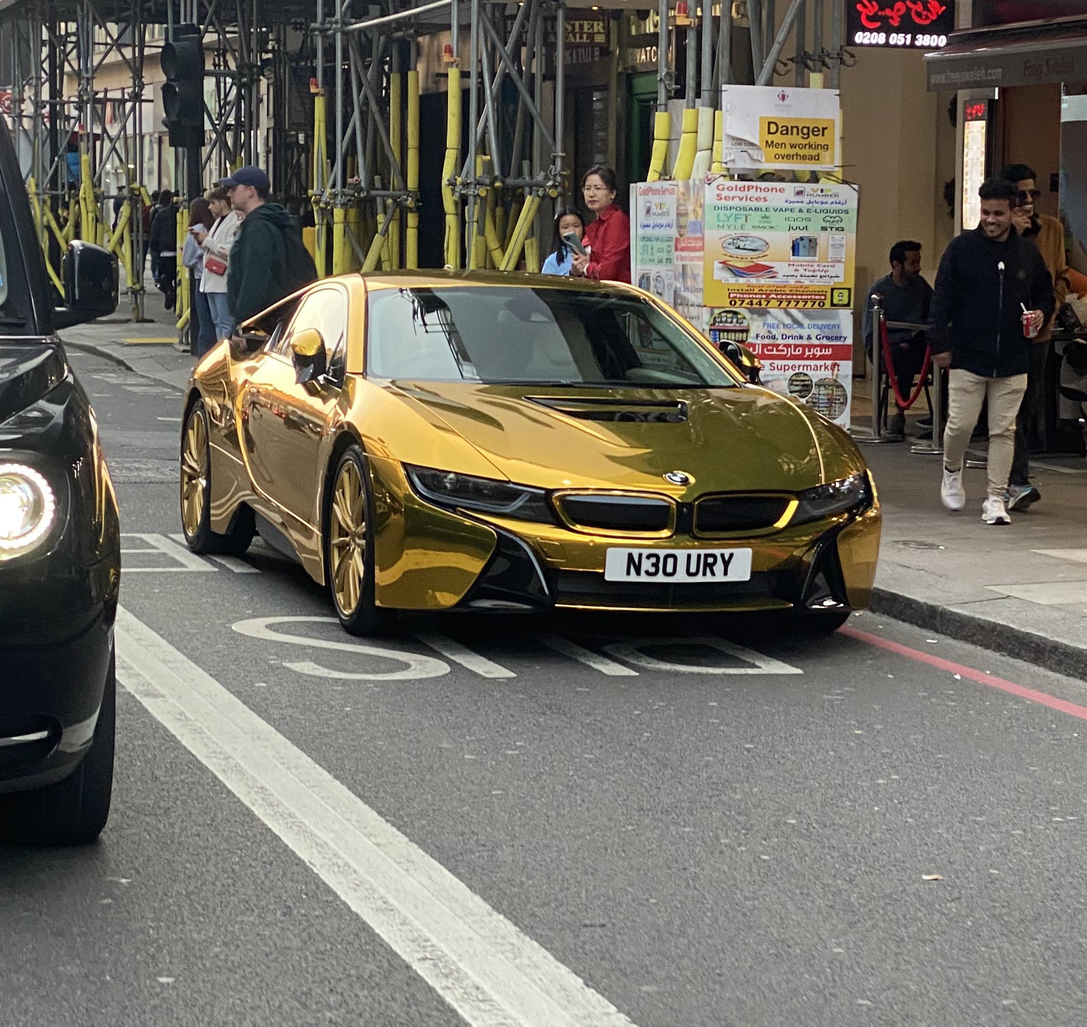 A shiny gold BMW sports car with the license plate "N30 URY" is parked on a city street near sidewalk diners and people walking by, with scaffolding and shops in the background.