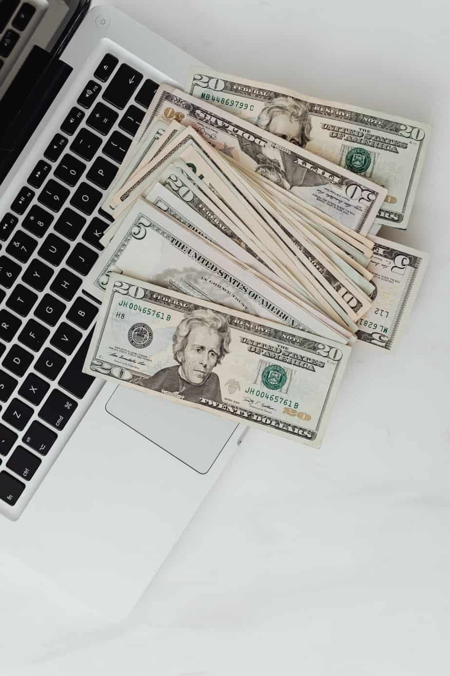 A stack of U.S. dollar bills, including $20 and $5 notes, spread across the keyboard of a silver laptop on a white surface.