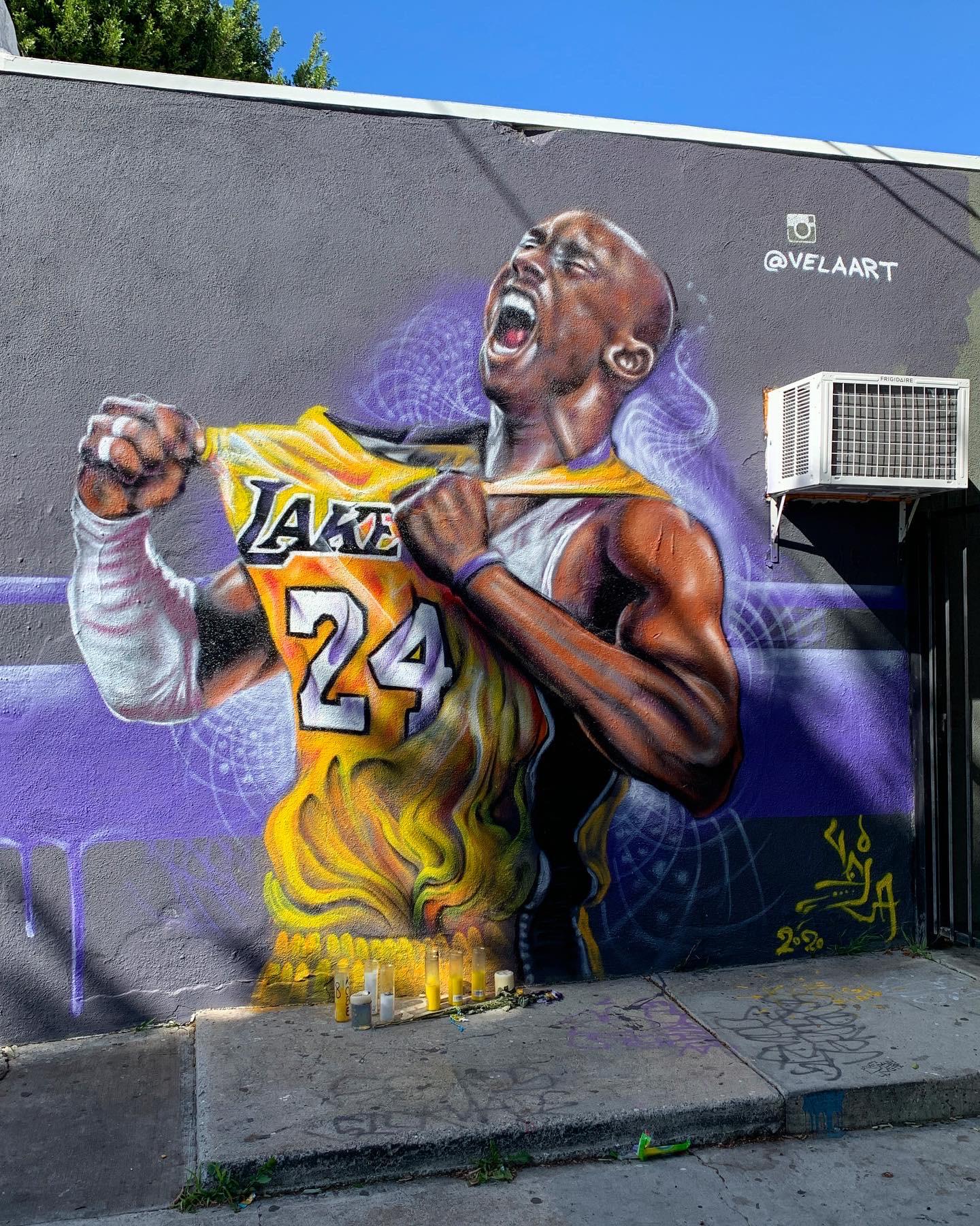 A mural on a wall depicts a basketball player in a yellow Lakers jersey, number 24, passionately pulling his jersey. Art candles are placed below the mural. The artwork includes the artist's handle "@VELAART" and an air conditioning unit.