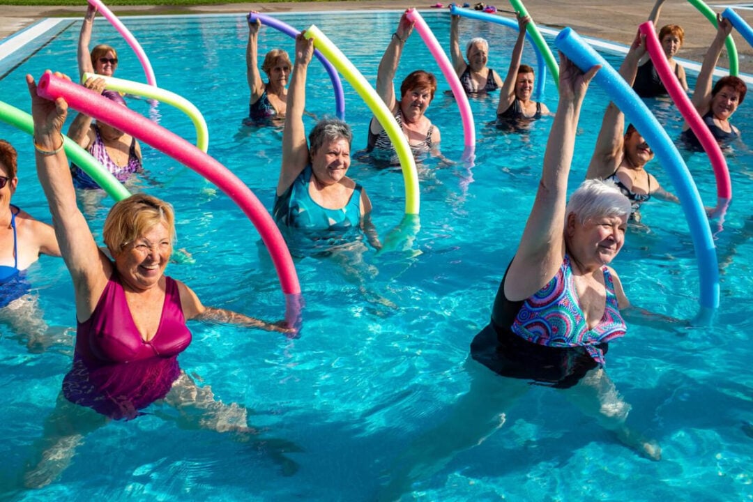 A group of older women in swimsuits participate in a water aerobics class in a pool, smiling and raising colorful foam pool noodles above their heads.