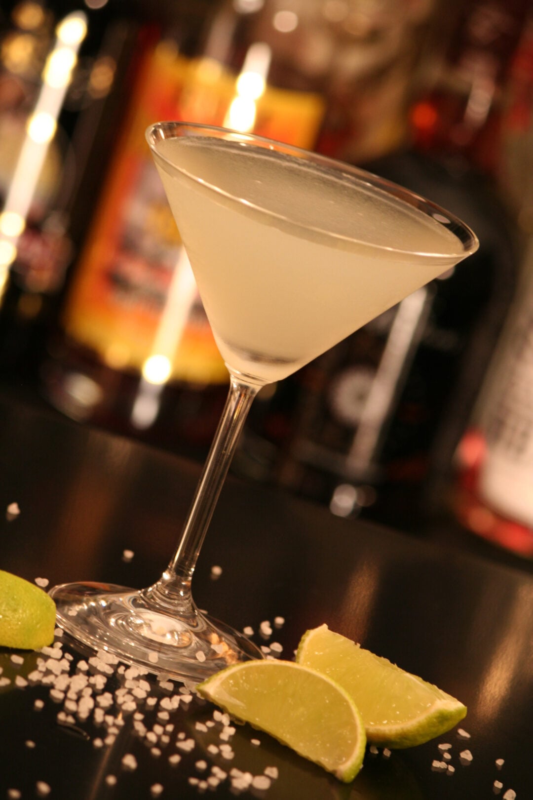 A classic cocktail in a martini glass with a light yellow drink, surrounded by lime wedges and scattered salt on a dark surface, with blurred bottles in the background.