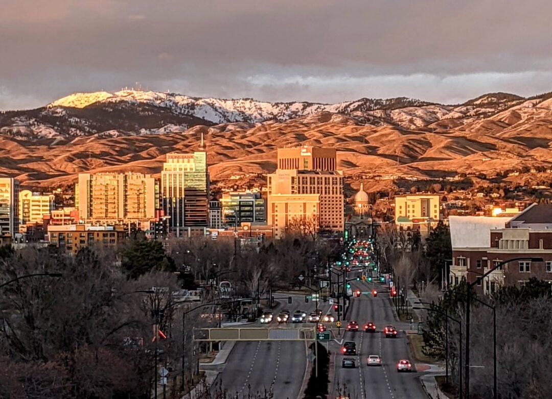 A cityscape at sunset shows tall buildings and a busy street with cars and traffic lights, with rolling golden hills and snow-capped mountains in the background.