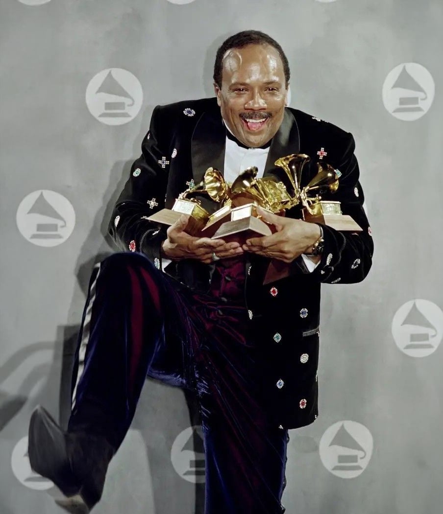 A joyful man in a black, decorated suit holds several Grammy Awards and lifts one leg in celebration, standing in front of a backdrop with repeating Grammy logos.