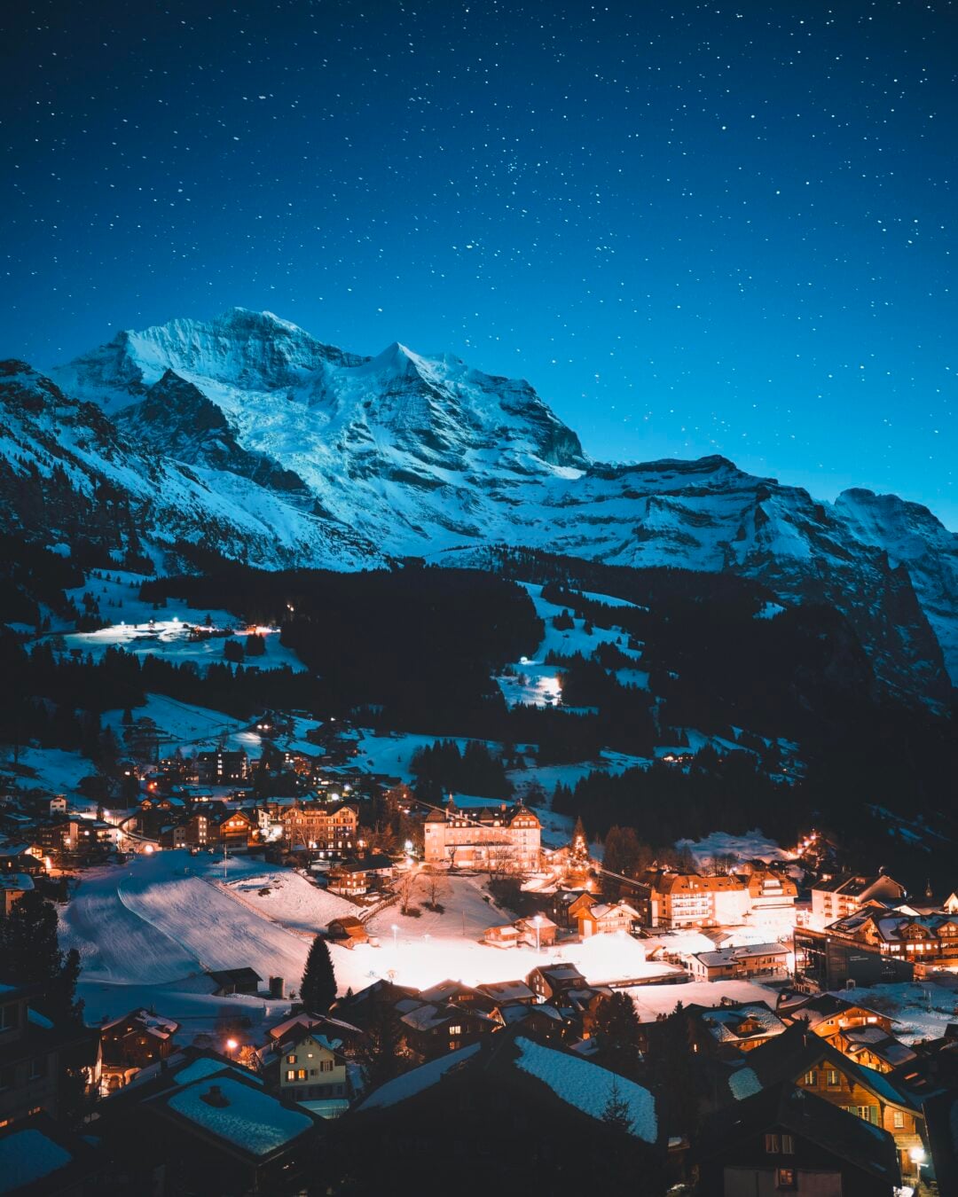 A snowy mountain village glows with warm lights under a clear, starry night sky, with majestic, snow-covered peaks rising in the background.