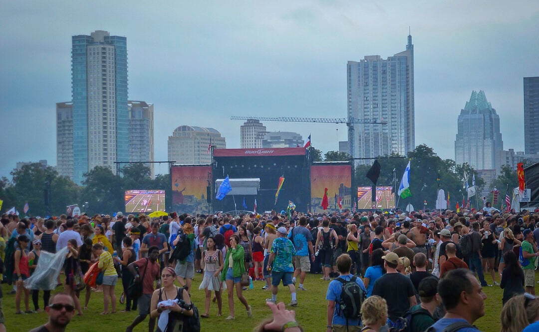 A large crowd gathers at an outdoor music festival with tall city buildings and a cloudy sky in the background. People are holding flags and standing in front of a stage, creating a lively atmosphere.