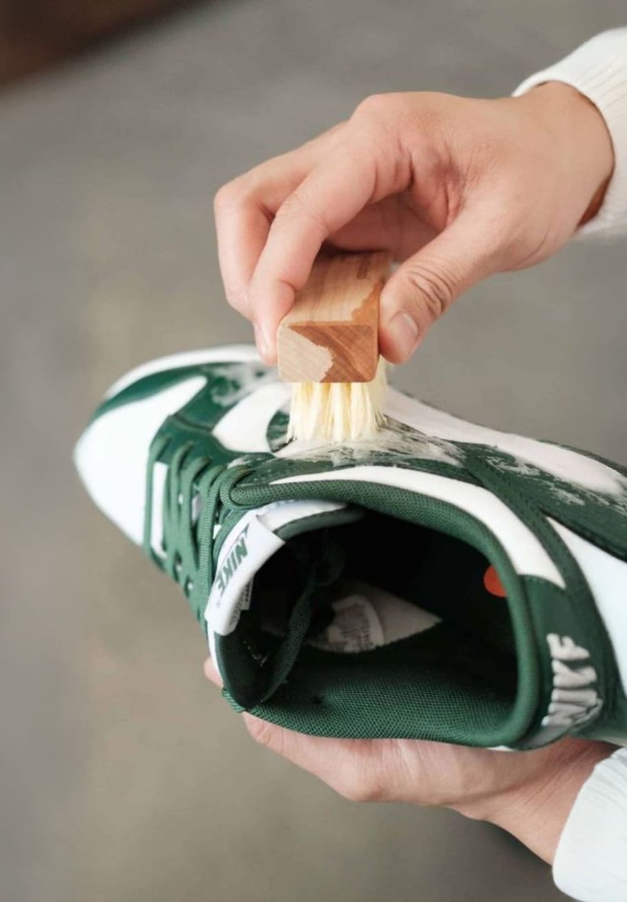 A person uses a small brush to clean the white and green upper part of a sneaker, applying soap or cleaning solution to remove dirt.