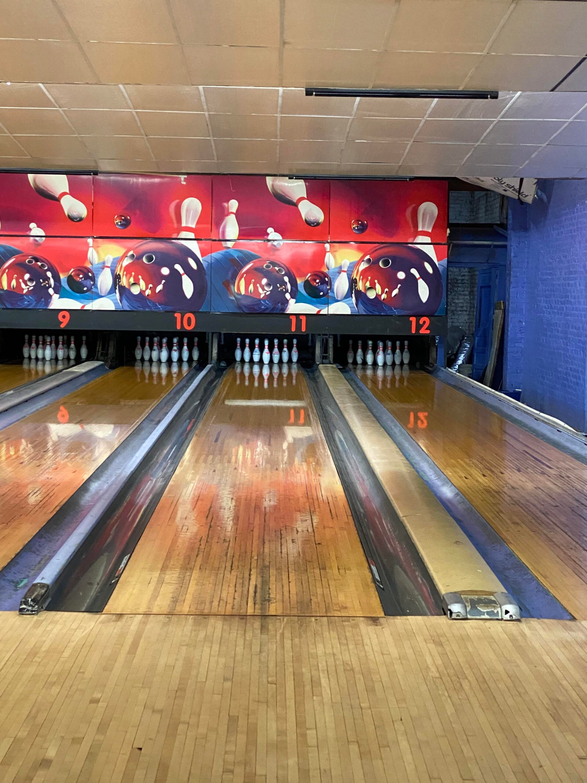 A bowling alley with four lanes numbered 9 to 12, each with ten white pins set up. The wooden floors are shiny, and a colorful mural of bowling balls and pins decorates the back wall.