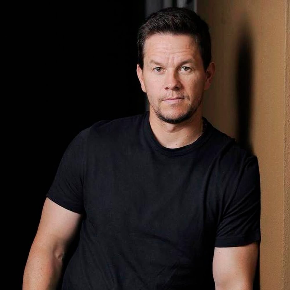 A man with short brown hair and a trimmed beard is wearing a plain black t-shirt, standing indoors against a tan wall and looking at the camera with a neutral expression.