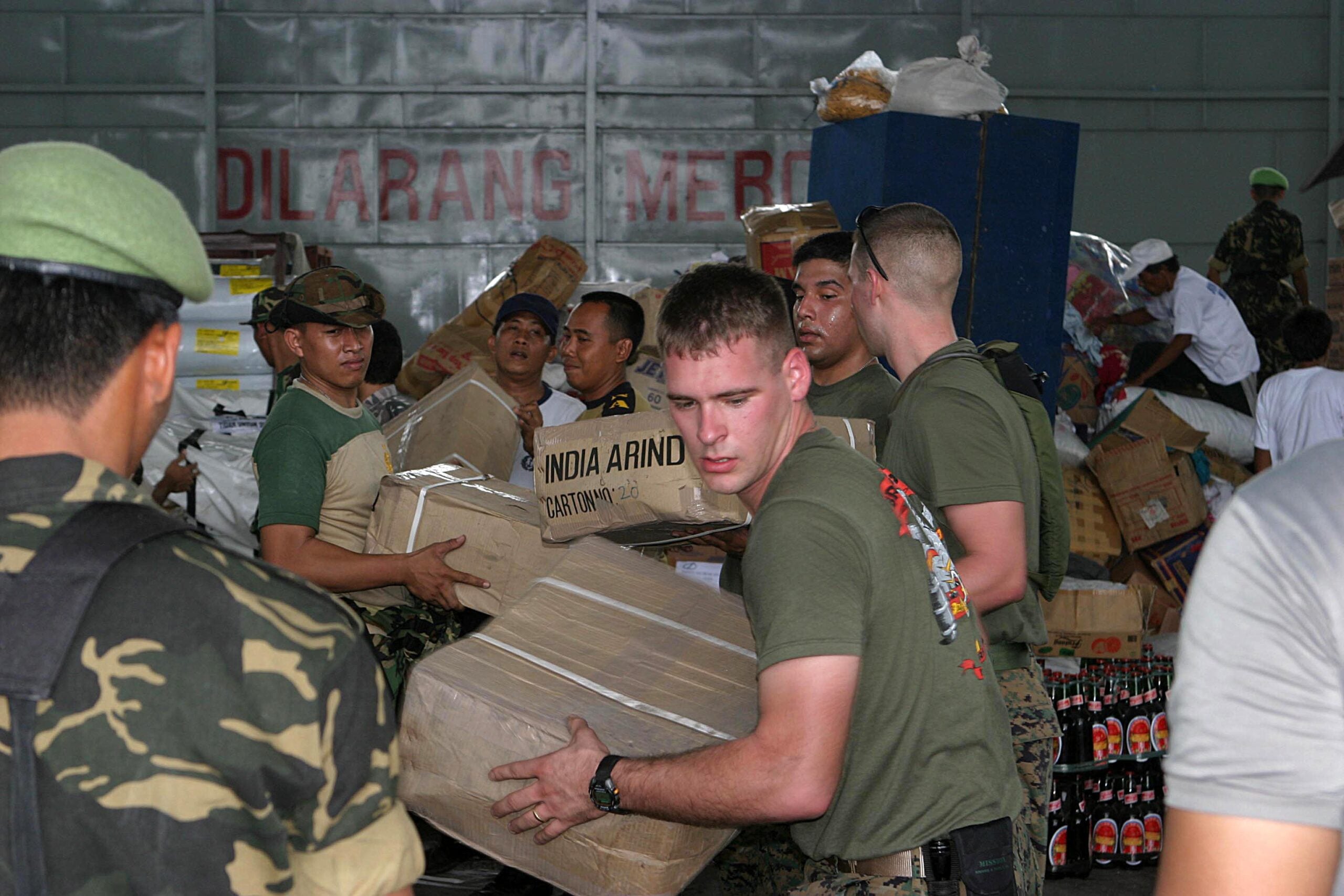 Several uniformed men work together to unload and carry large cardboard boxes filled with supplies inside a warehouse; stacks of goods and bottled drinks are visible in the background.