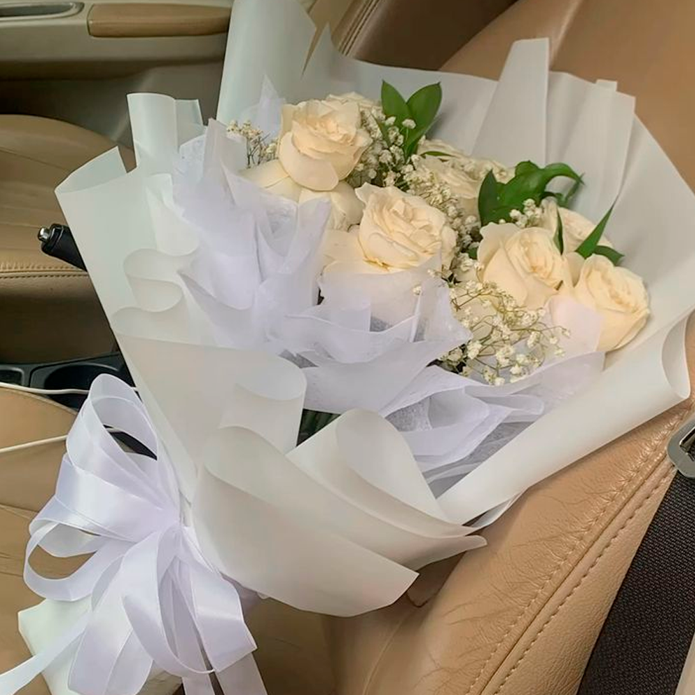 A bouquet of pale cream roses, wrapped in white paper with baby's breath and greenery, sits on a tan leather car seat. The bouquet is tied with a large white ribbon bow.