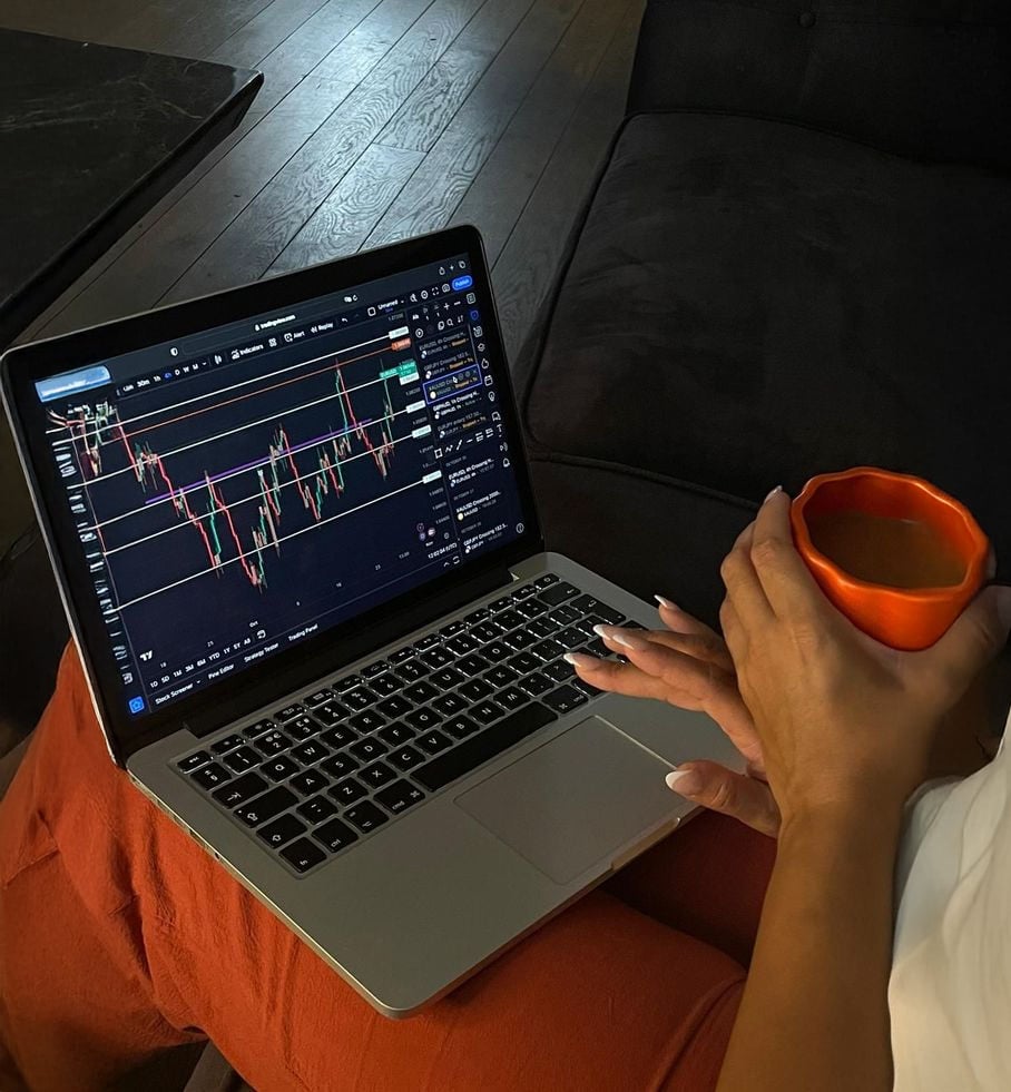 A person wearing orange pants holds an orange mug of coffee while analyzing a candlestick stock chart on a laptop, sitting on a dark sofa with wooden flooring visible.