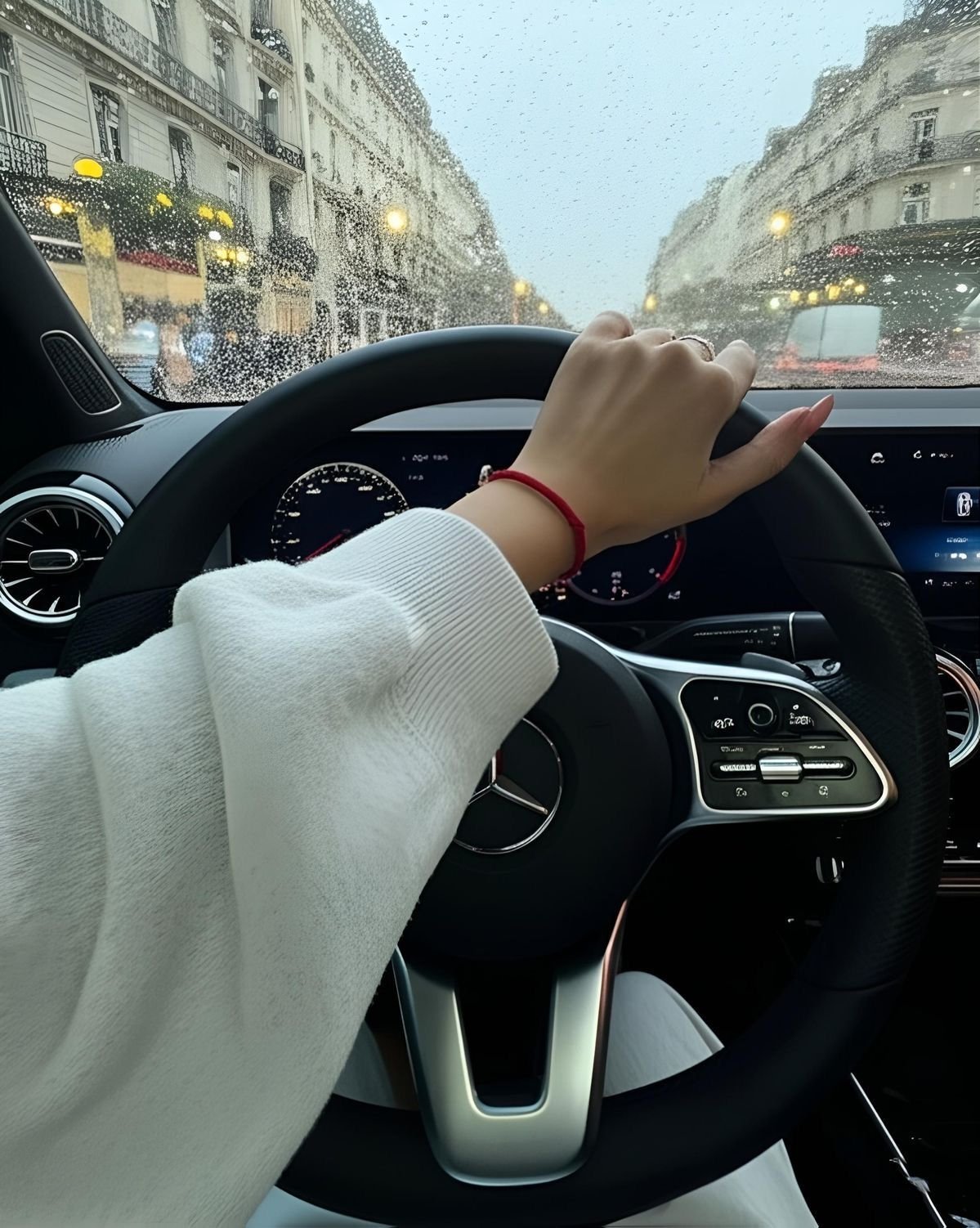 A person wearing a white sleeve and red bracelet is holding the steering wheel of a Mercedes-Benz. Through the windshield, a rainy city street with classic buildings and streetlights is visible.