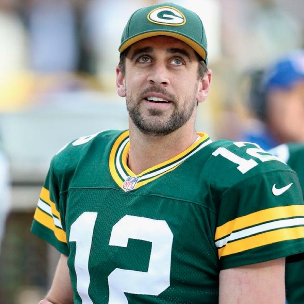 A football player in a green and yellow Green Bay Packers uniform and cap, wearing jersey number 12, looks upwards with a slight smile on a bright day.