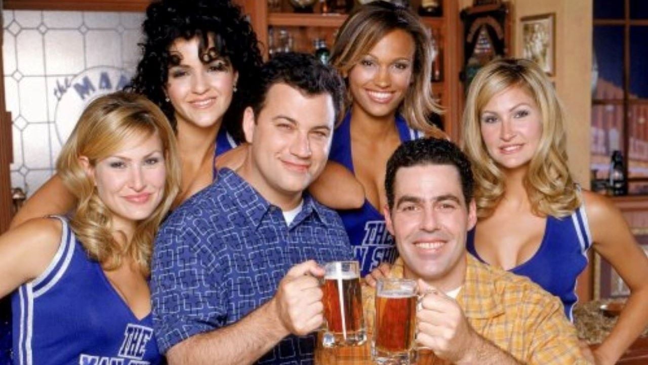 Two men holding beer mugs smile at the camera, surrounded by four women in matching blue cheerleader-style outfits, all posing together in a bar-like setting.