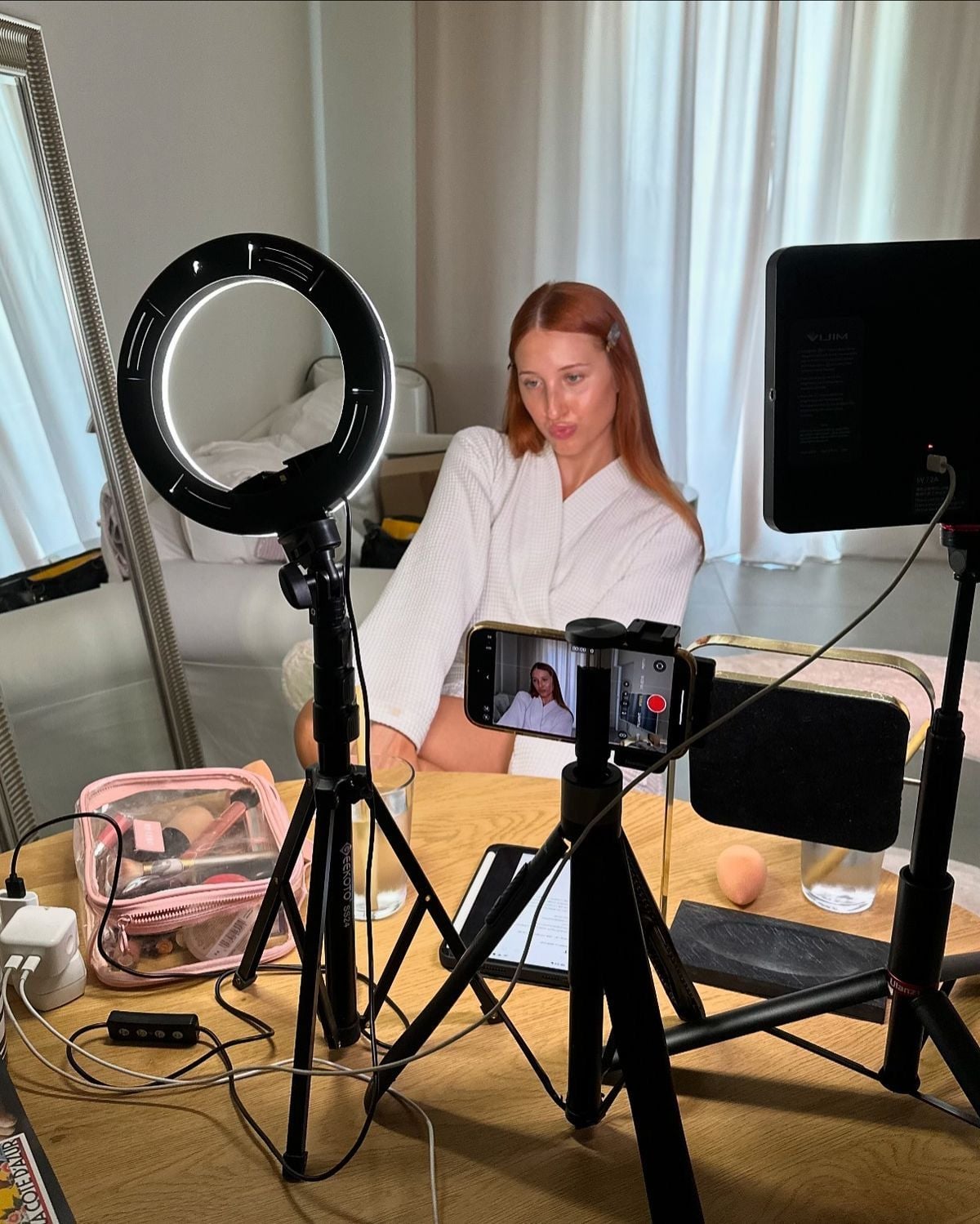 A woman with long red hair and a white robe sits near a table with tripods, ring light, camera, and makeup items, preparing to film or livestream content in a bright room with white curtains.