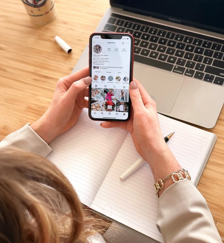 A person holds a smartphone displaying an Instagram profile while sitting at a desk with an open laptop, a blank notebook, a pen, and a cup of coffee.