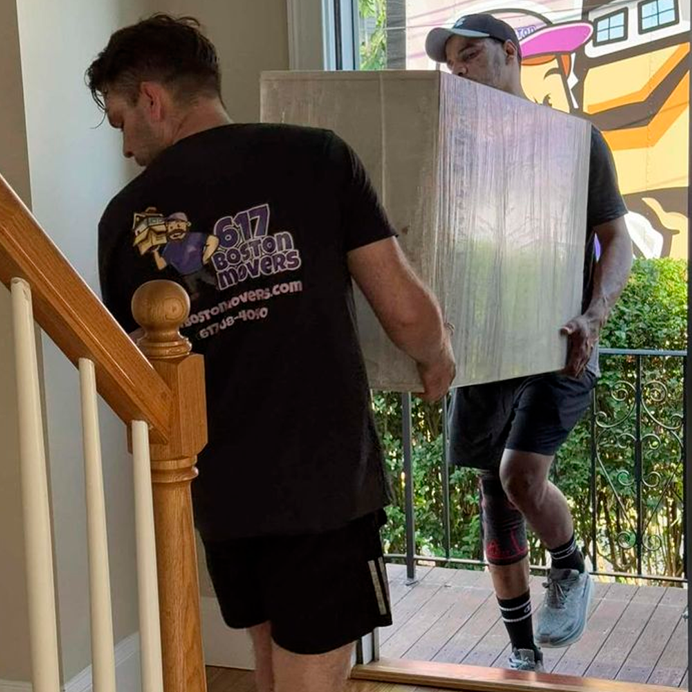 Two movers carry a large, wrapped box up a staircase inside a house. One mover wears a “617 Boston Movers” t-shirt, and sunlight streams in through a doorway with a mural visible outside.