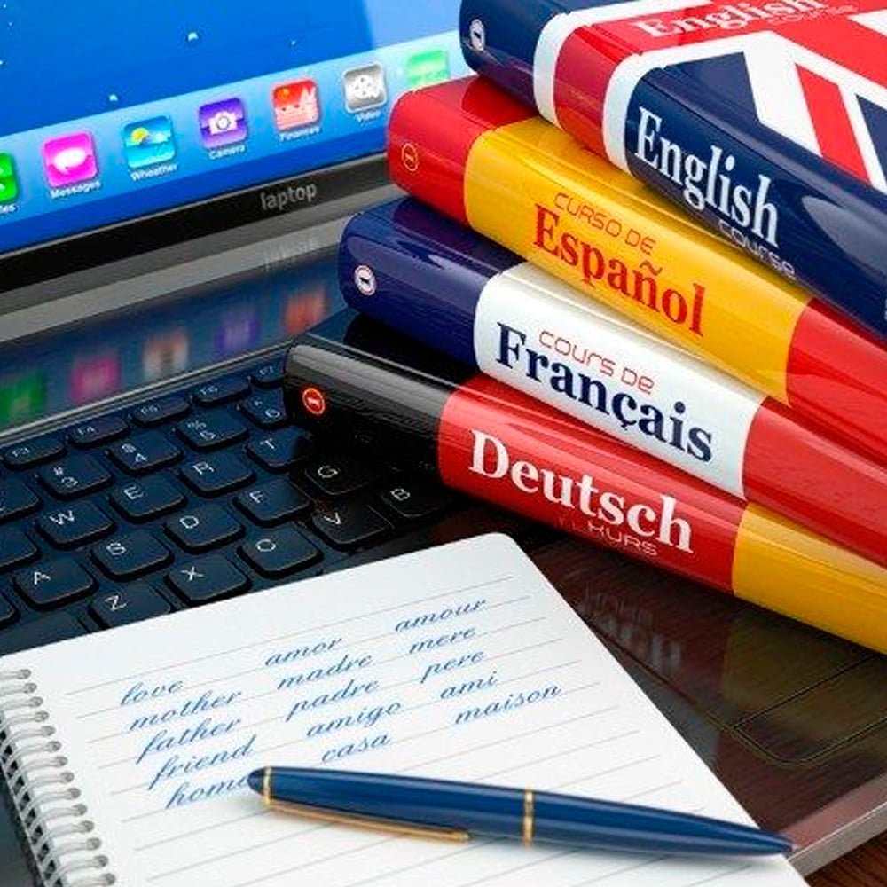 A laptop with a stack of language books—English, Spanish, French, and German—next to a notepad showing English words and their translated equivalents, with a pen resting on the notepad.