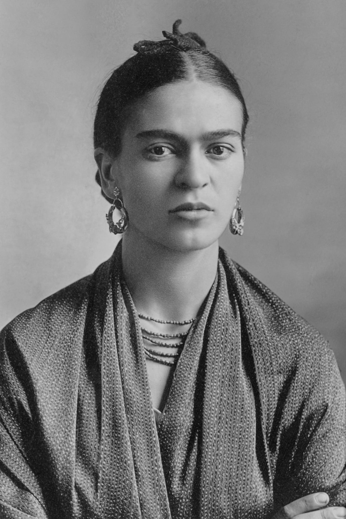 Black and white portrait of a woman with dark hair tied back, wearing large earrings, layered necklaces, and a textured shawl draped over her shoulders, looking directly at the camera with a neutral expression.