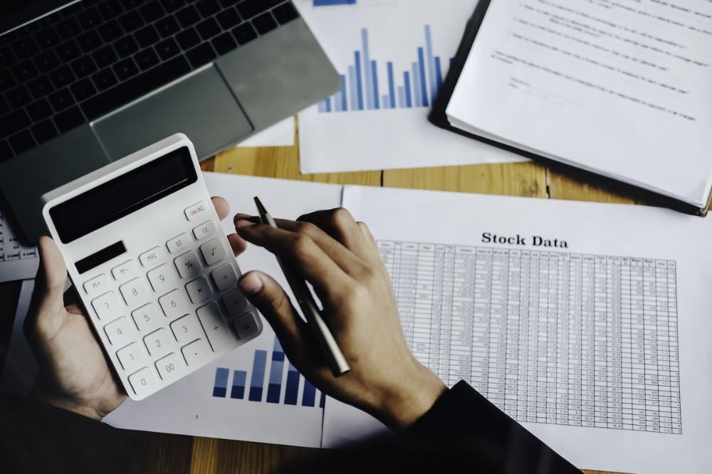Person using a calculator with a pen in hand, sitting at a desk with stock data sheets, charts, documents, and a laptop. The scene suggests financial analysis or business calculations.