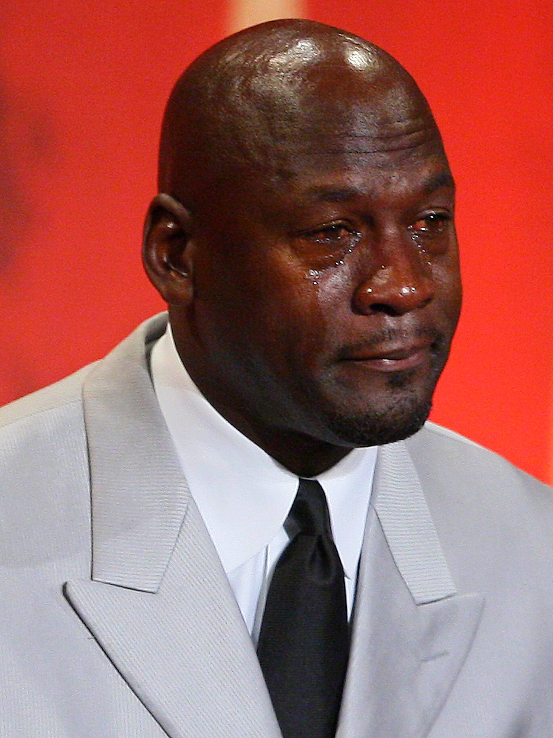 A man in a light gray suit and black tie is visibly emotional, with tears streaming down his face, set against a red background.
