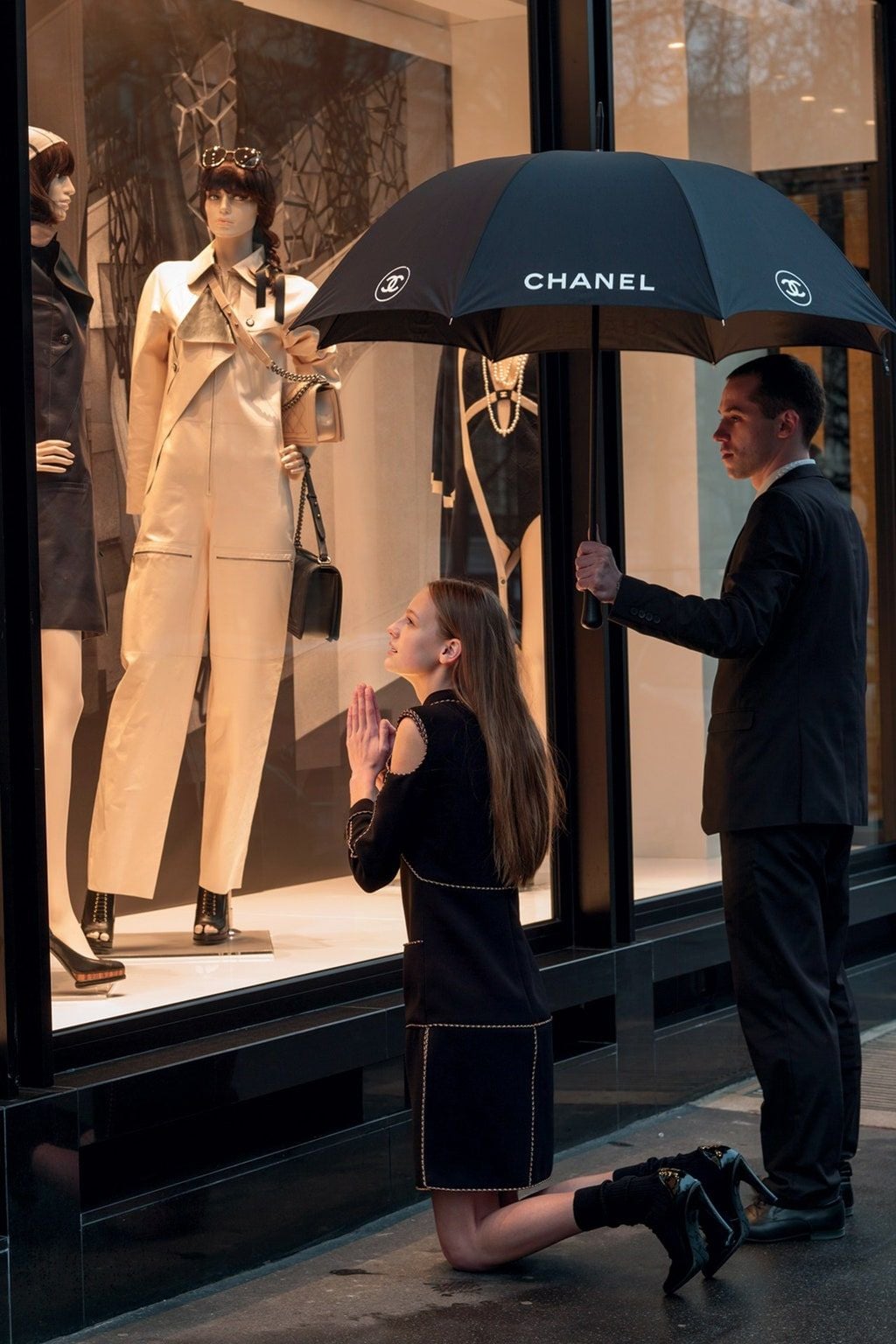 A woman kneels on the sidewalk, hands clasped in prayer, gazing at mannequins in a Chanel store window. A man beside her holds a large black Chanel umbrella over her, shielding her from rain.