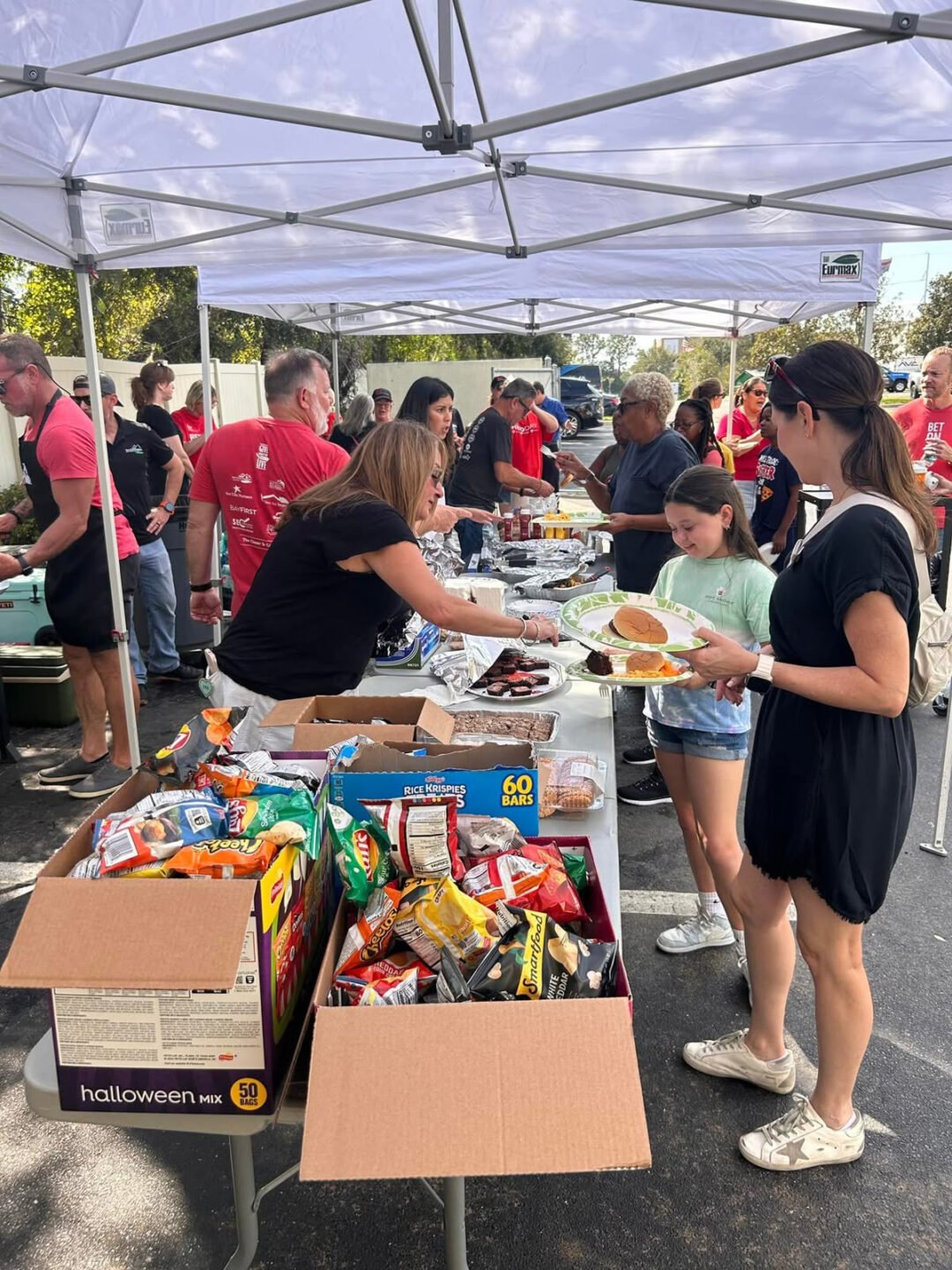 People gather under canopy tents at an outdoor event, serving and receiving food from tables filled with snacks, chips, drinks, and various dishes. A woman and a girl hold plates at the front of the line.