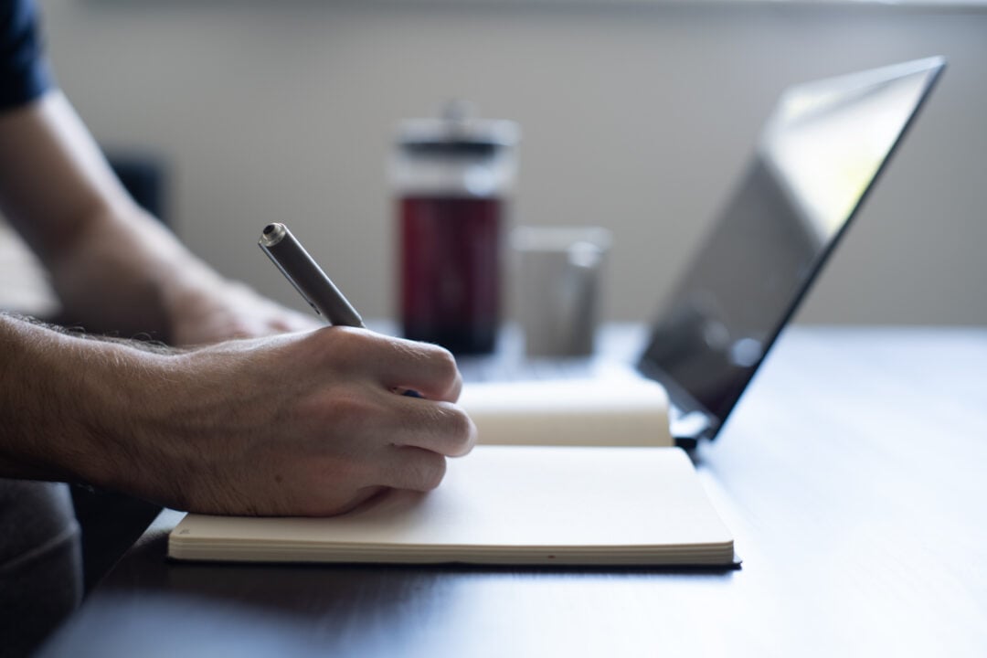 A person writes in a notebook with a pen at a desk, next to an open laptop and a French press coffee maker, with soft natural light coming from a nearby window.