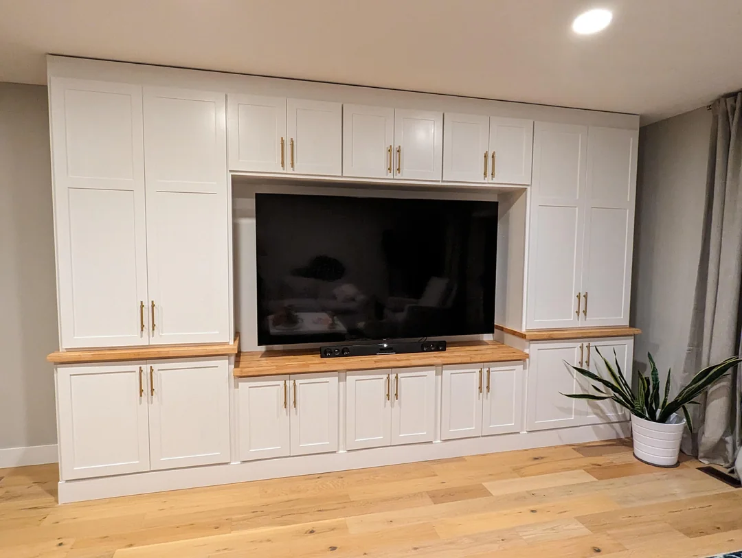 A large flat-screen TV is centered in a built-in white entertainment unit with cabinets and gold handles, set against a light-colored wall. A potted plant sits on the wooden floor to the right.
