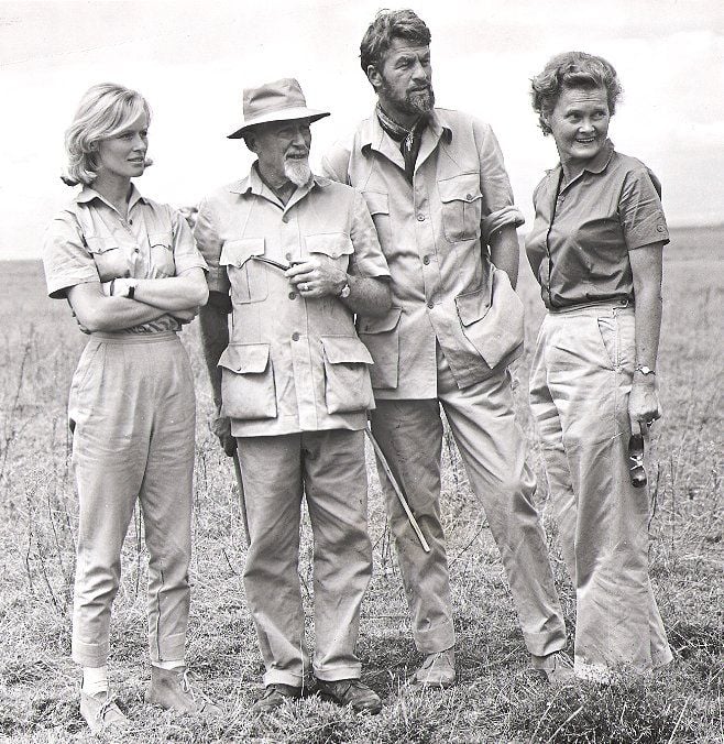 Four people stand together outdoors in a grassy field, dressed in safari-style clothing. Two men and two women appear to be posing and talking, with a relaxed and casual demeanor. The photo is in black and white.