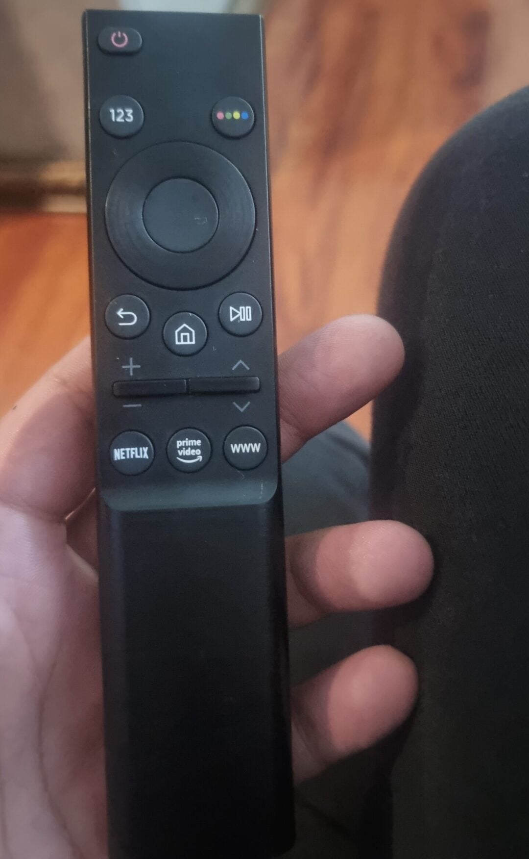 A hand holds a black TV remote control with round directional buttons, volume controls, and shortcut buttons for Netflix, Prime Video, and a WWW browser. A couch and wood floor are visible in the background.