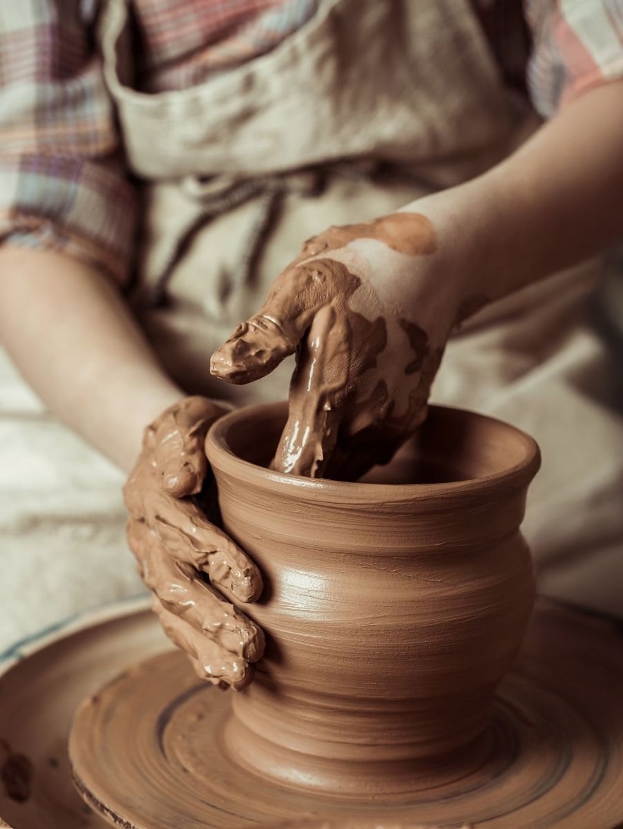 A person shapes a clay pot on a spinning pottery wheel with both hands, wearing a beige apron and a plaid shirt. Their hands are covered in wet clay as they mold the vessel.