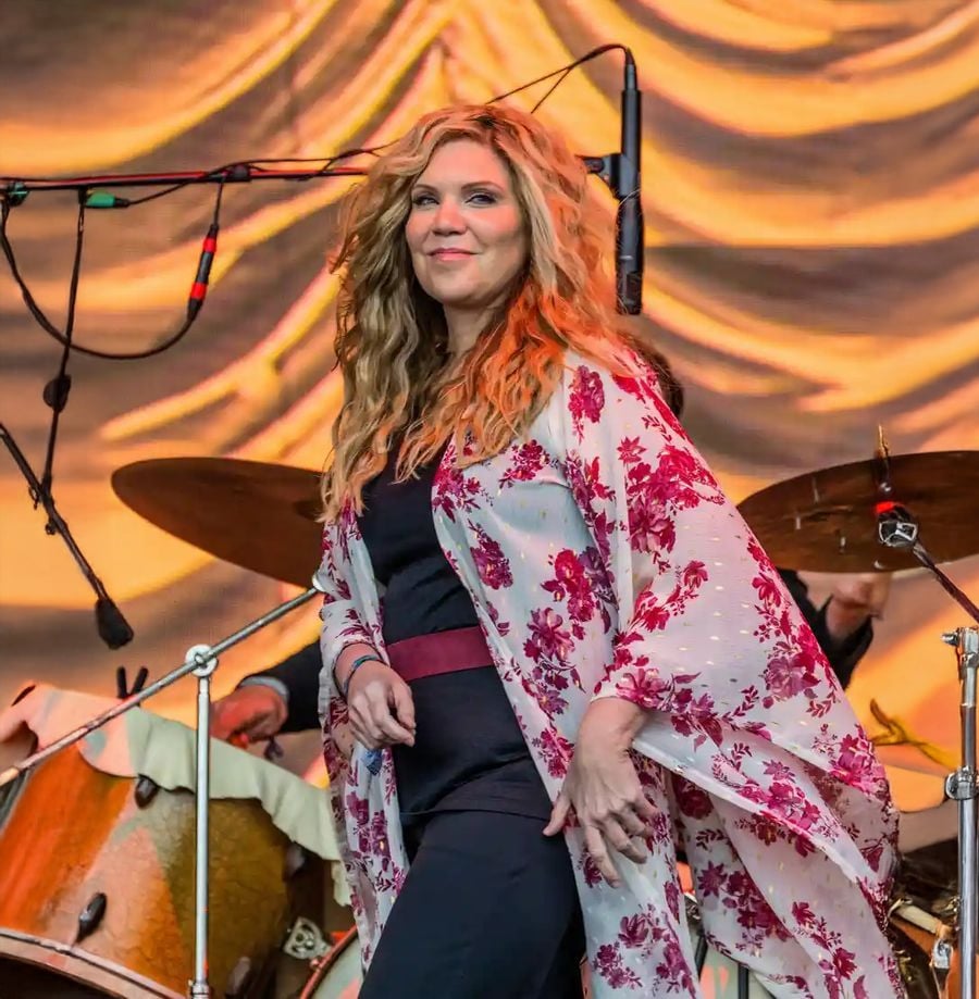 A woman with long, wavy hair wears a floral kimono over a black outfit while standing on stage in front of a drum set and a gold, draped backdrop, with microphones overhead.