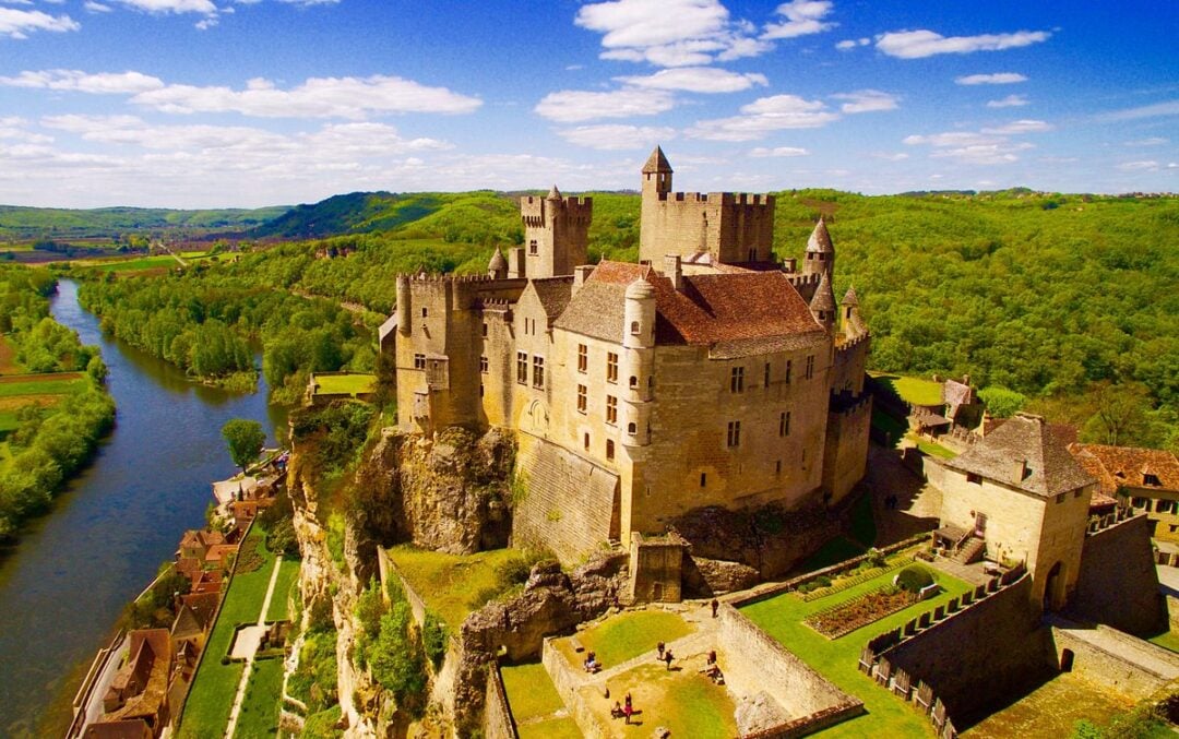 A large medieval stone castle sits atop a cliff overlooking a winding river and green forested landscape, with blue skies and scattered clouds above.
