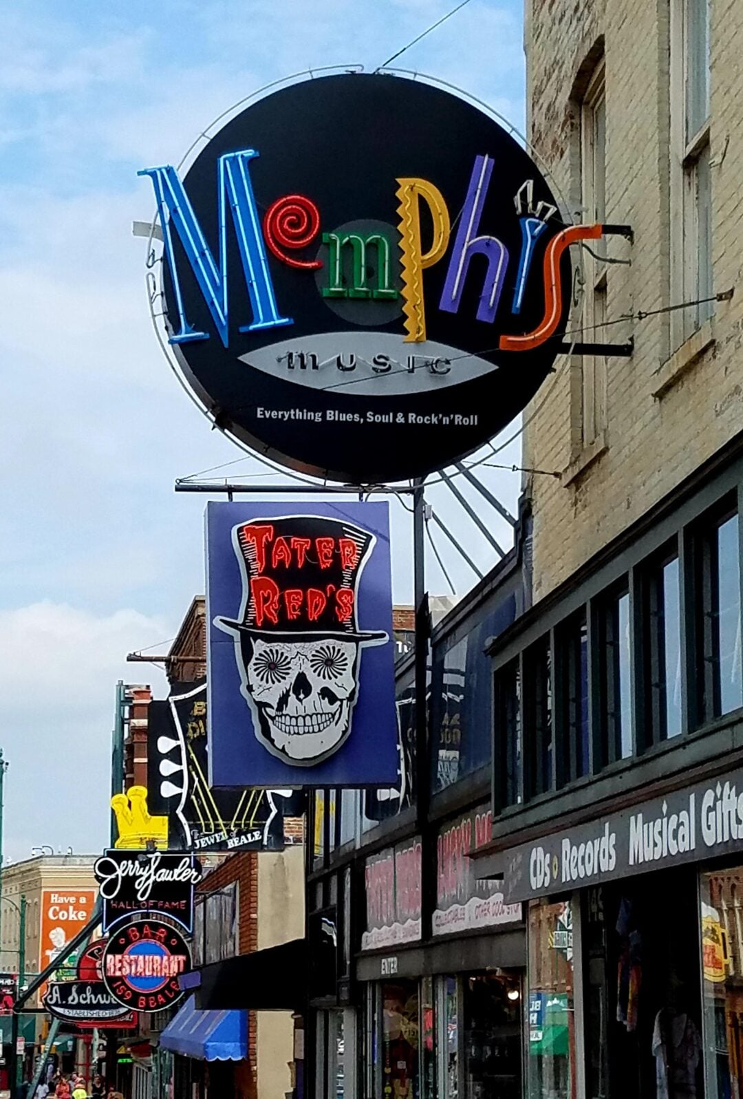 Colorful neon signs hang above a street in Memphis, including a large "Memphis Music" sign and a "Tater Reds" sign with a skull in a hat. Other storefronts and signs are visible along the busy street.