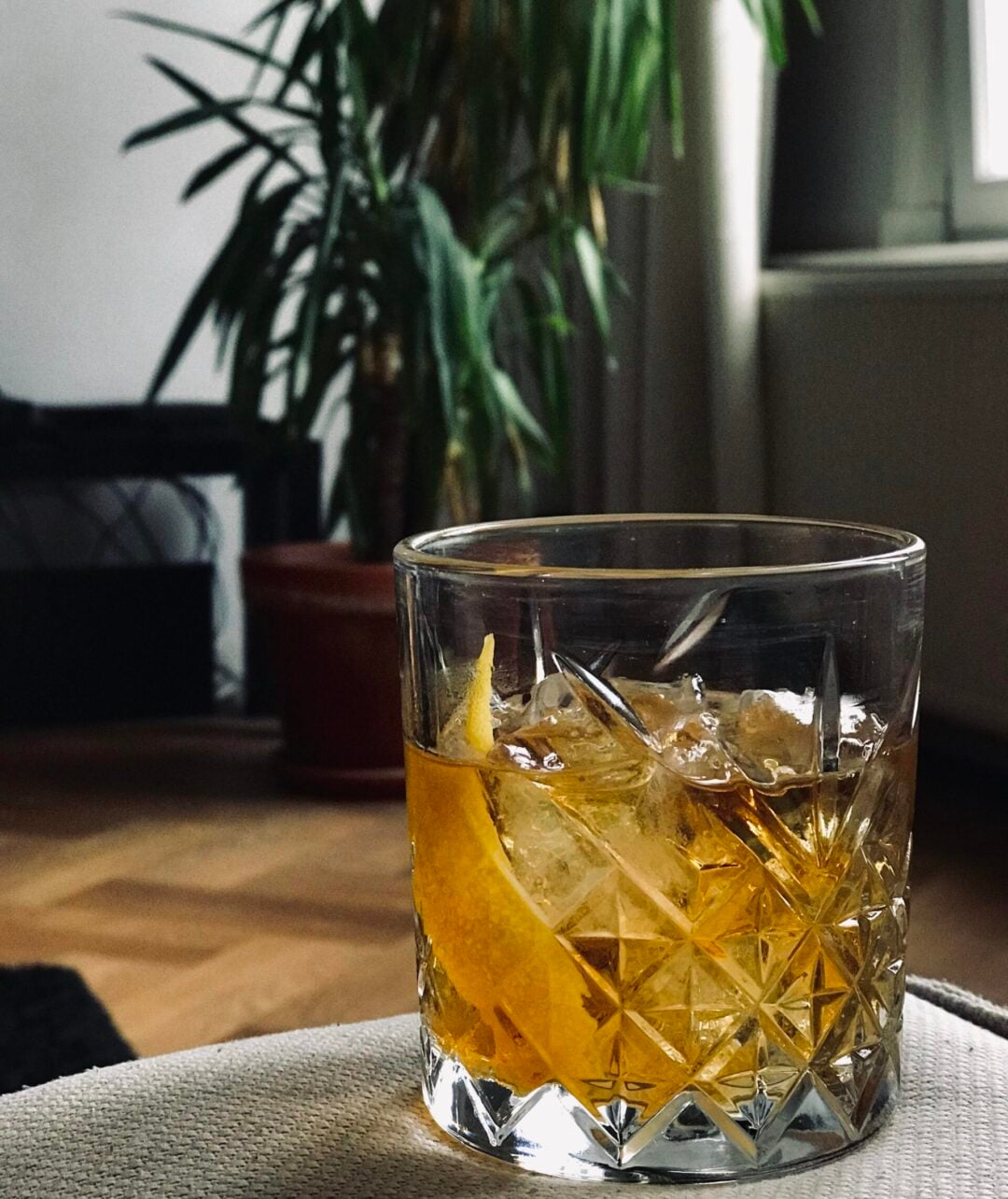 A crystal glass filled with an amber cocktail, ice cubes, and a lemon twist sits on a light-colored surface. In the background, a potted plant and a window are visible in a cozy indoor setting.