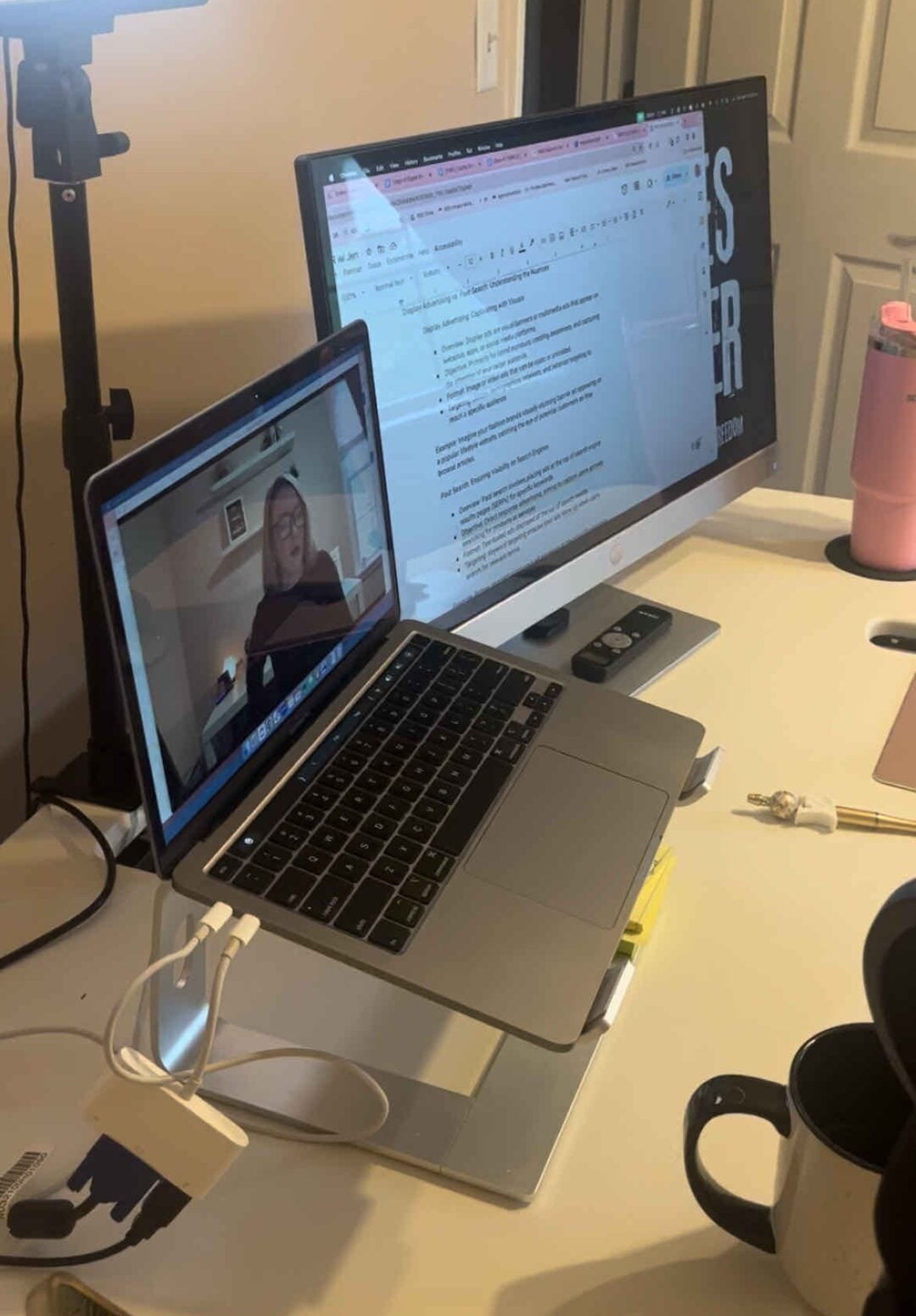 A workspace with a laptop showing a woman on a video call, a large monitor displaying a document, a water bottle, a mug, and a ring light on a stand. The desk is organized with various office items.