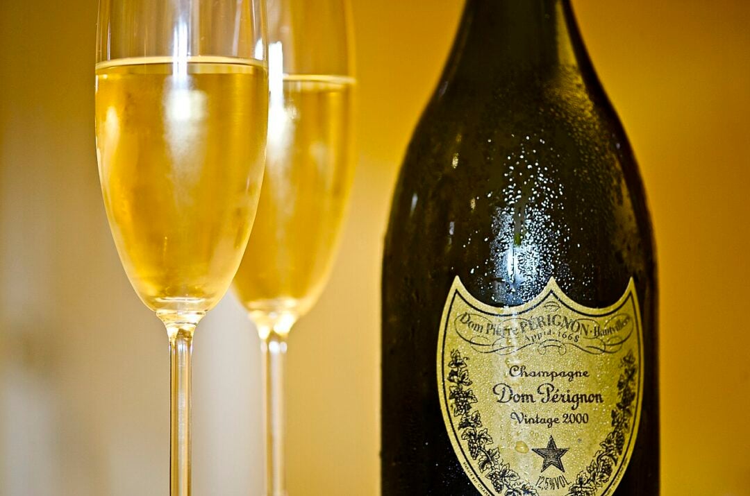 Two filled champagne flutes stand beside a chilled bottle of Dom Pérignon Vintage 2000, with condensation on the bottle and a warm yellow background.