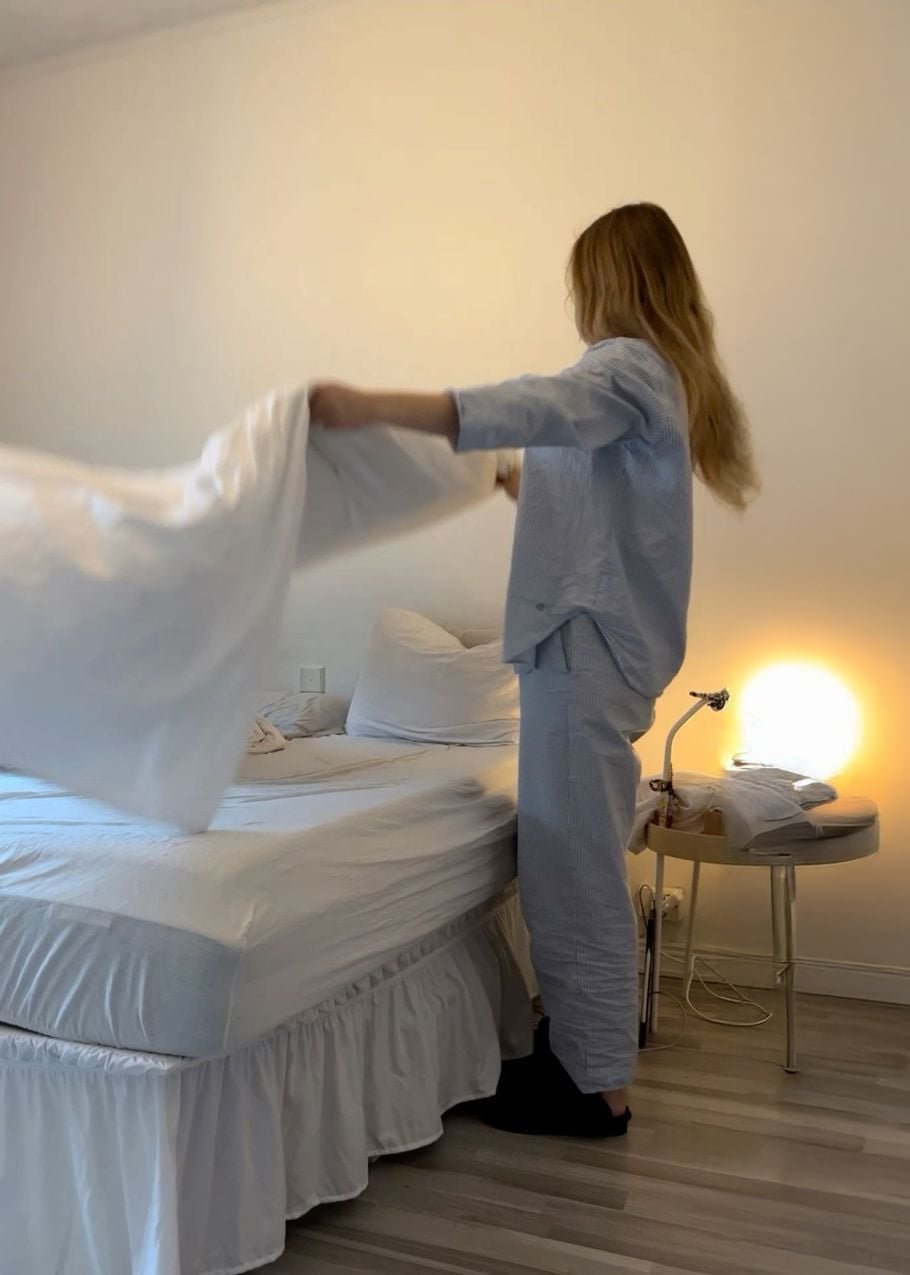 A person in pajamas is making a bed, spreading a white sheet. The room has light-colored walls, a bedside table with a lamp, and folded clothes. The floor is light wood.